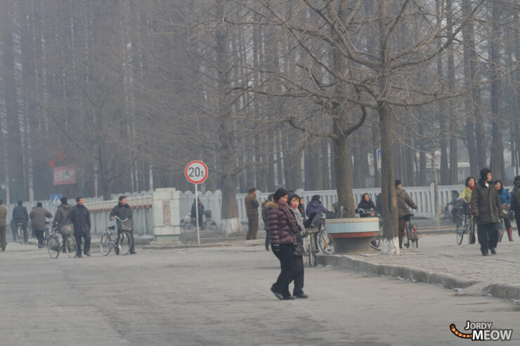 Everyday life scene in Pyongyang, North Korea: people walking, cyclists, urban setting, overcast sky.