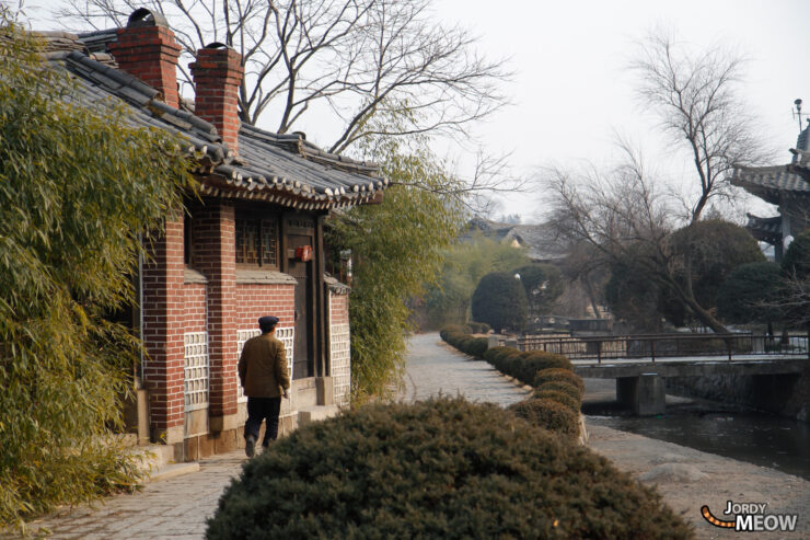 Tranquil North Korean village scene with traditional building, lush greenery, and peaceful waterway.