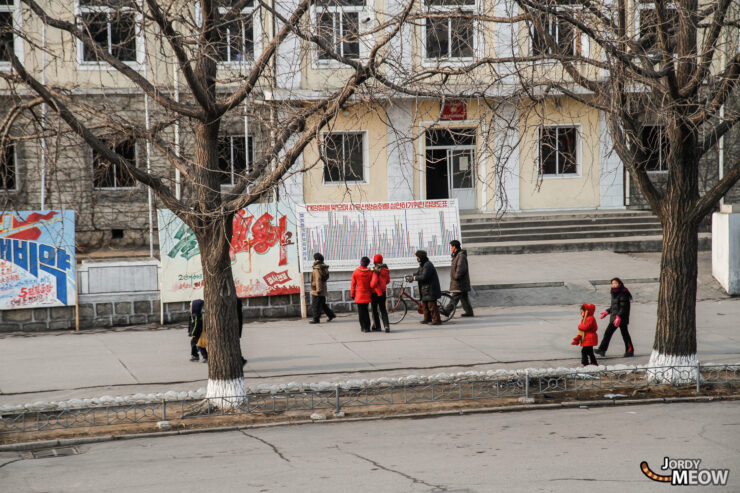 People in red jackets on sidewalk in North Korea urban scene with institutional building.