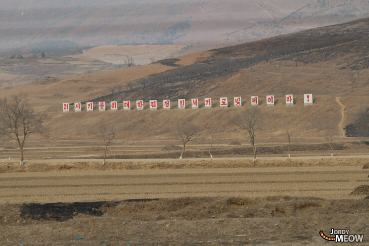 Exploring rural North Korea with scenic hills, barren fields, and political red flags.
