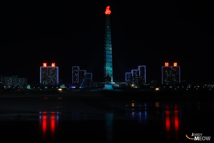 Pyongyang skyline at night with illuminated buildings and tower reflecting in still waters.
