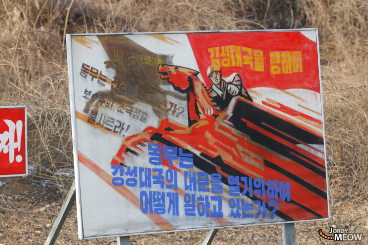 North Korea propaganda poster: soldier in action with red flag and patriotic text.