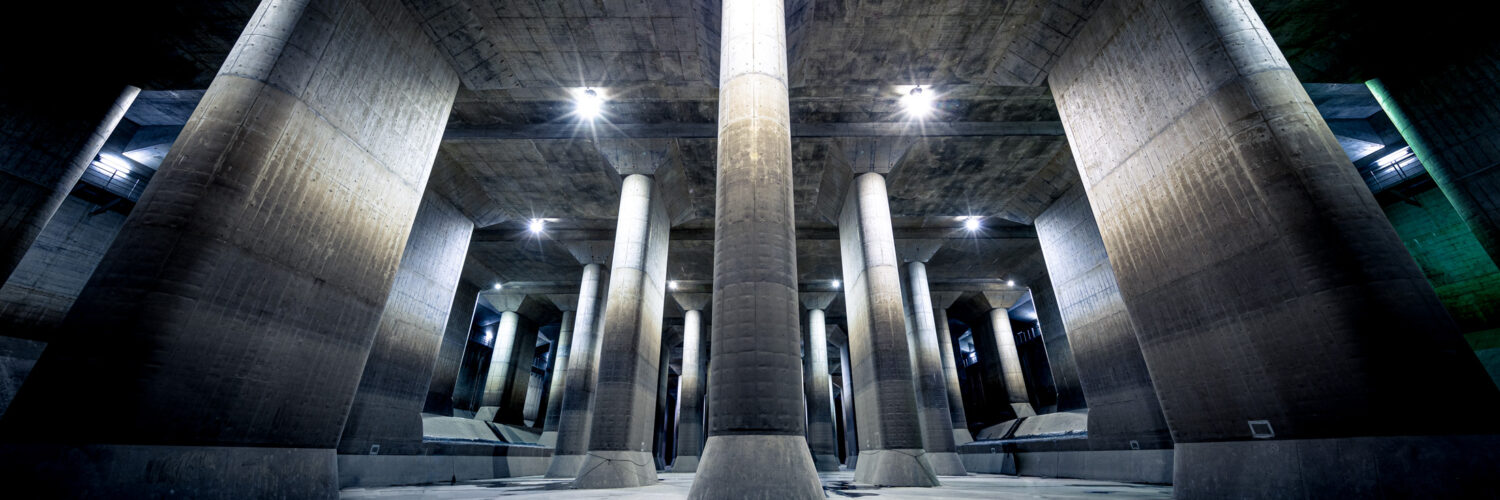 Subterranean flood water diversion channel with towering concrete columns and dramatic lighting.